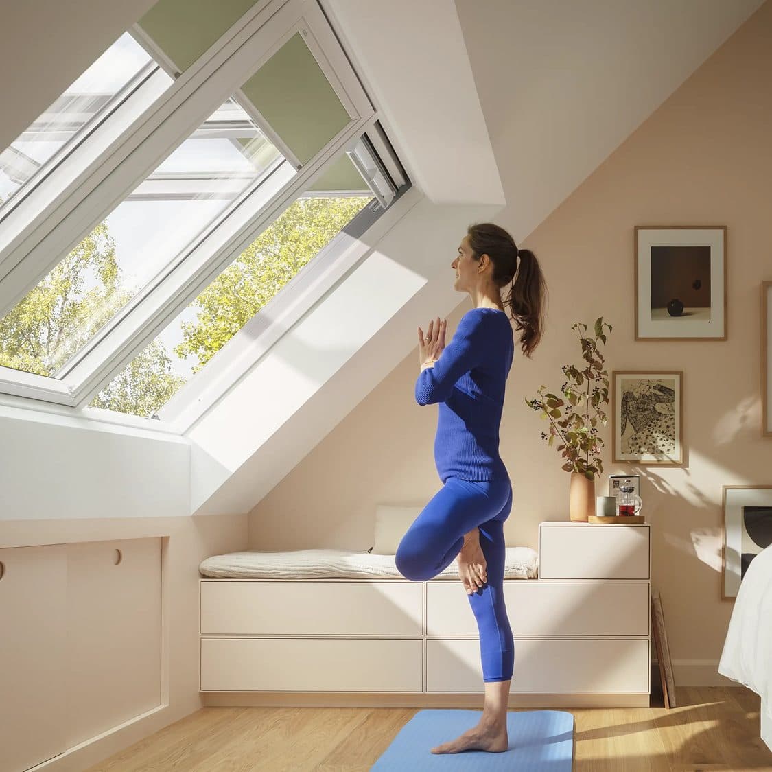 A woman doing yoga on a mat in a bright attic room, one of the two roof windows above her propped open.