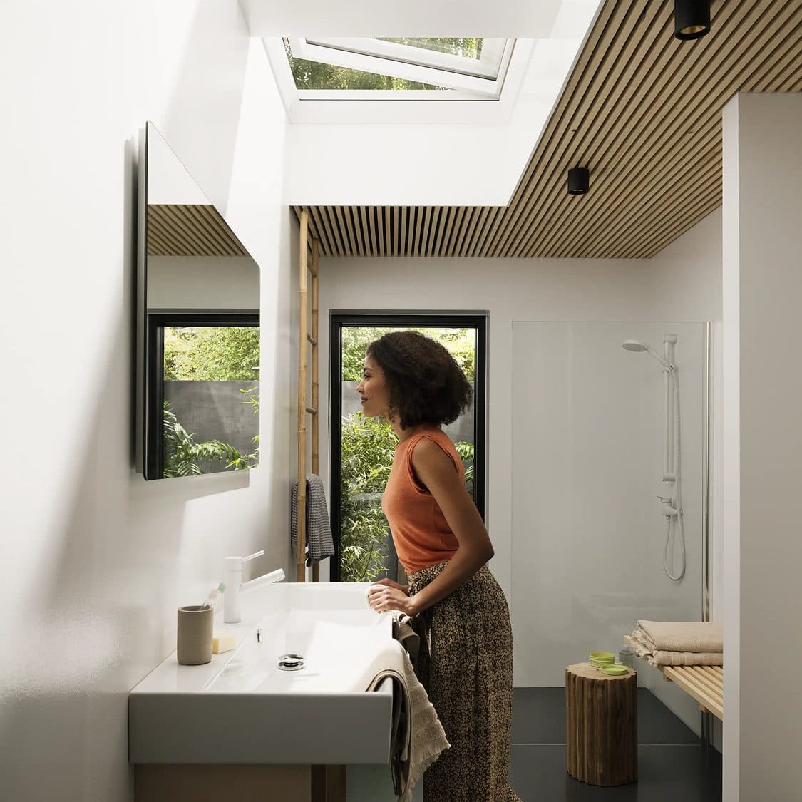 A woman at the basin of a bathroom under a roof window, a shower to one side and a window onto greenery to the other.