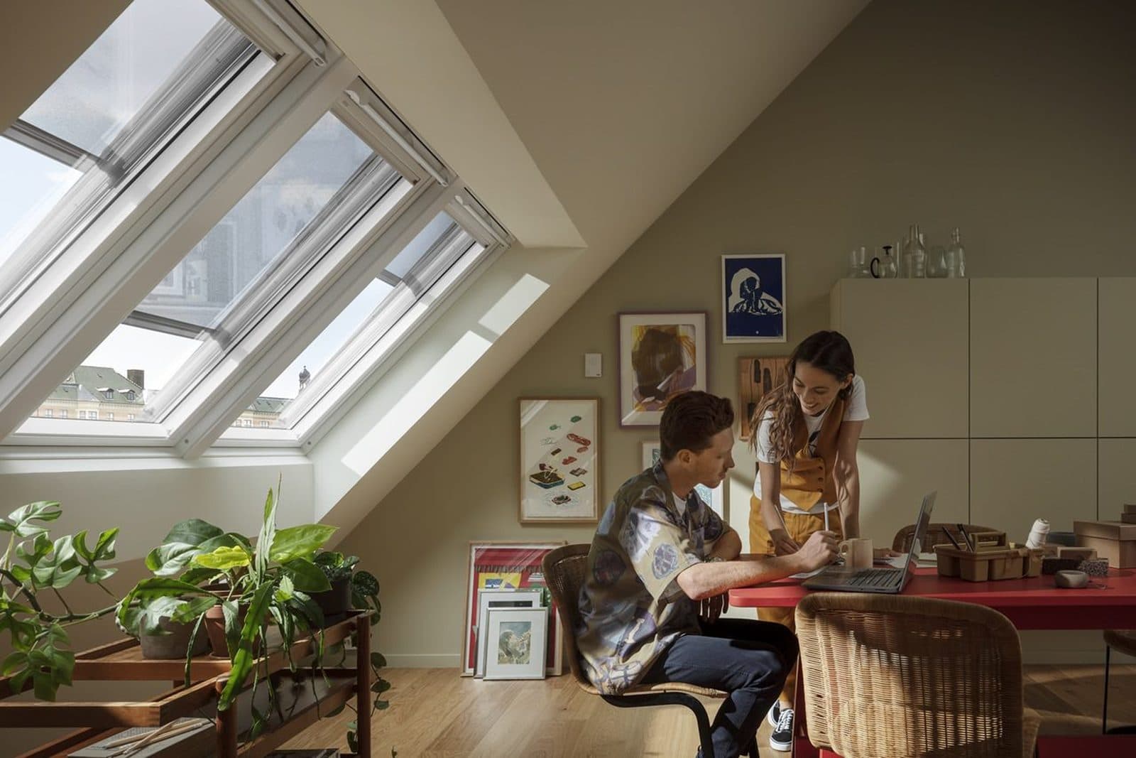 Two people working together at a desk in an attic room, under a run of large roof windows with plants and framed prints along the wall.